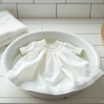 Robe bébé baptême en dentelle blanche posée à plat pour être lavée à la main dans un bassin en céramique avec du savon doux